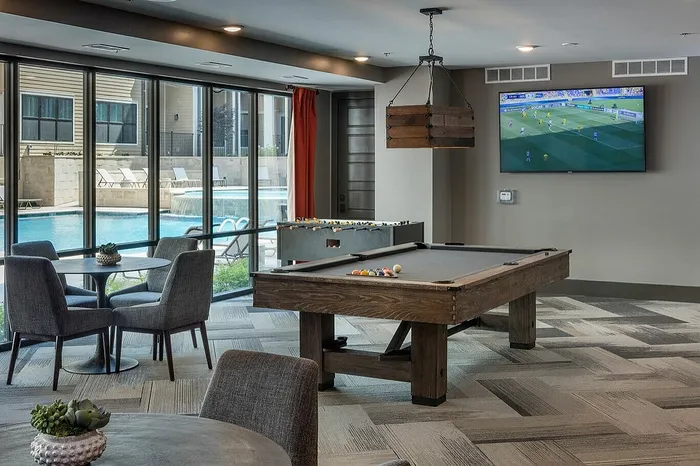 Luxurious entertainment room with a pool table, foosball, modern seating, and expansive pool views through large glass windows.