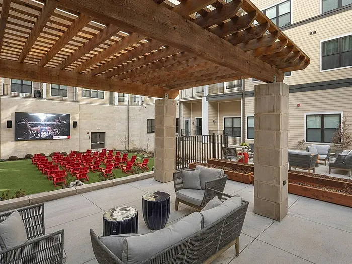 Modern apartment courtyard featuring cozy outdoor seating, vibrant red chairs on a lush lawn, and a large TV screen for communal entertainment under a stylish pergola.