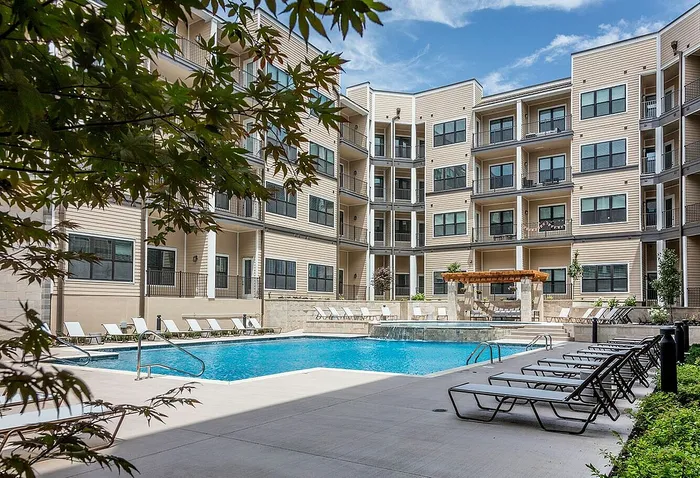 Modern apartment complex showcasing a luxurious pool area surrounded by stylish lounge chairs and lush greenery under a clear blue sky.
