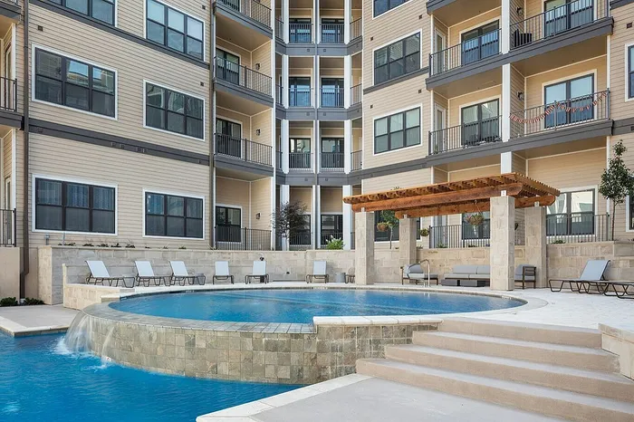 Luxurious apartment complex showcasing a sparkling pool with a cascading waterfall feature, elegant lounging area with pergola, and modern balcony views.