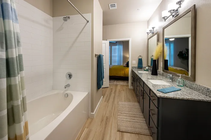 Elegant bathroom with bright lighting, featuring a luxurious bathtub, modern dual-sink vanity with granite countertop, and a glimpse of a cozy bedroom with a yellow accent.