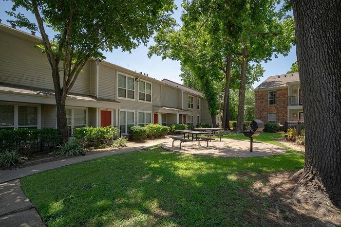 Charming apartment exterior featuring lush green landscaping, inviting picnic area, and mature trees for shade and tranquility.
