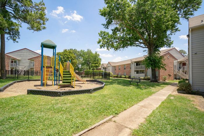 Charming apartment complex with lush greenery, featuring a vibrant playground under a clear blue sky, ideal for family living.