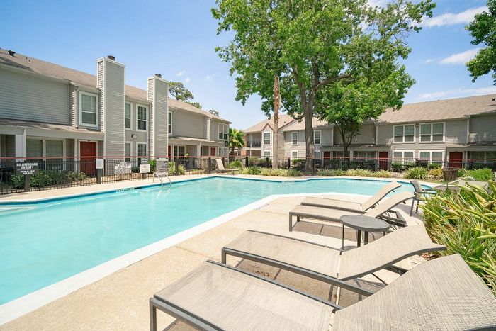 Inviting apartment community featuring a sparkling swimming pool with comfortable lounge chairs, set against charming two-story buildings and lush greenery.