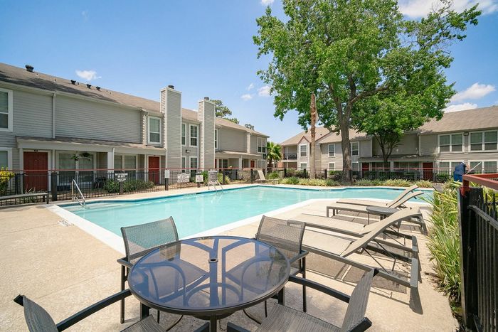 Inviting apartment community pool area with lounge chairs, patio seating, and lush greenery on a sunny day.