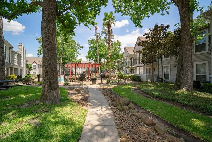 Charming apartment community with lush green landscaping, a shaded pathway, and inviting outdoor grilling area under a pergola.
