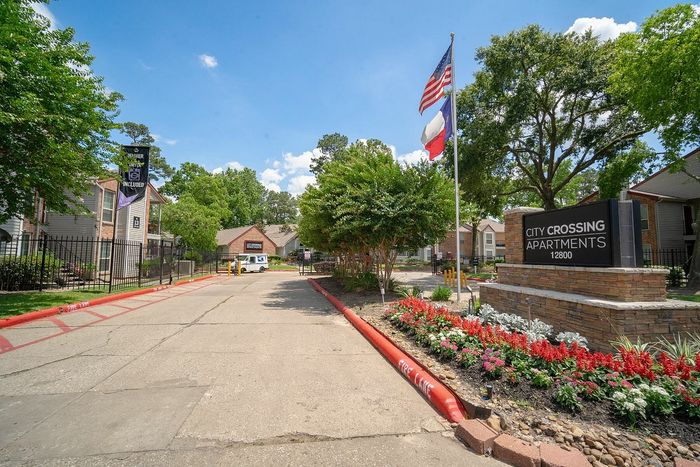 Inviting entrance to City Crossing Apartments with vibrant landscaping, waving flags, and a welcoming street view under a clear blue sky.