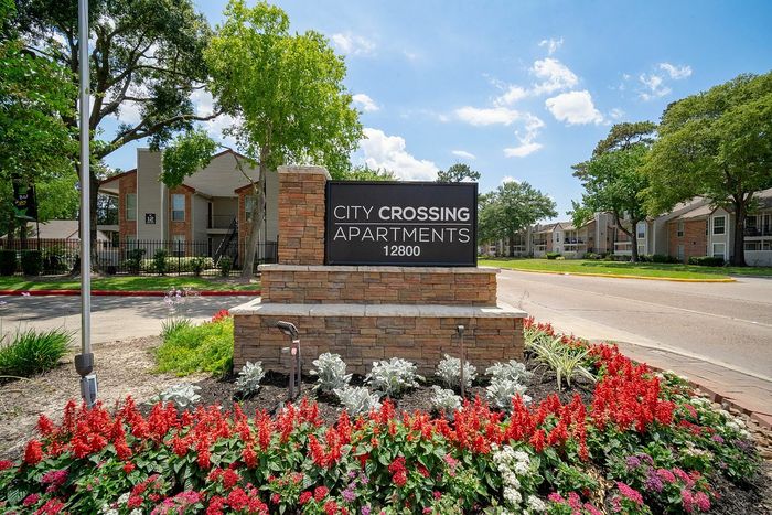 Inviting entrance to City Crossing Apartments surrounded by vibrant flowers and lush greenery on a bright, sunny day.