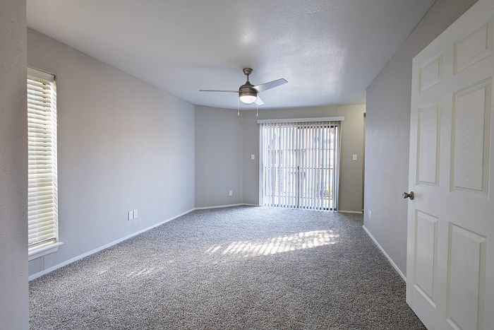 Bright and spacious living room with modern ceiling fan and plush carpeting, featuring large windows and sliding glass doors leading to a sunny outdoor space.