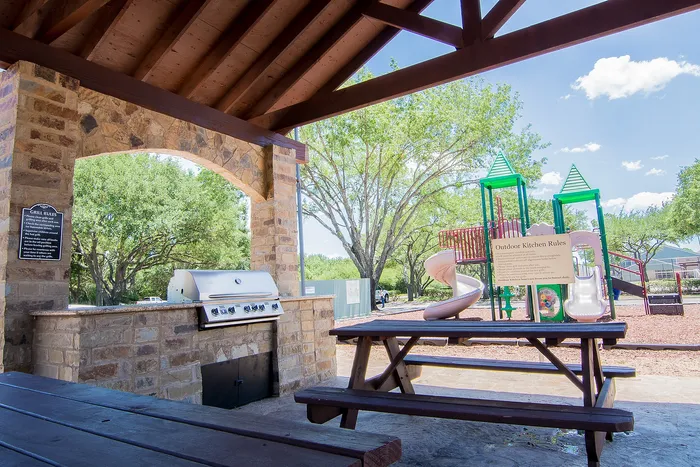 Charming outdoor space featuring a brick grill station and a cozy picnic table under a wooden pavilion, with a vibrant playground and lush greenery in the background.