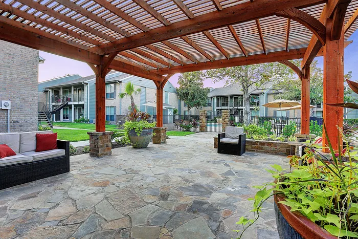 Charming outdoor patio with stone flooring, cozy seating, and lush greenery under a stylish wooden pergola, perfect for relaxation.
