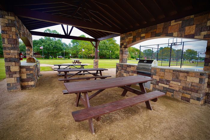 Charming outdoor pavilion with rustic stone walls and picnic benches, offering a perfect setting for gatherings. Includes a modern grill and views of lush green spaces and a basketball court.