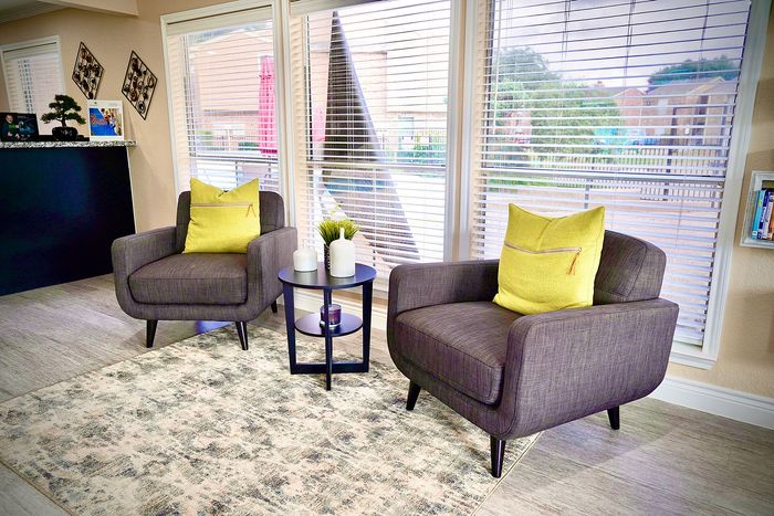 Bright and inviting seating area with modern gray armchairs, vibrant yellow cushions, and a stylish coffee table, framed by large windows offering sunny natural light.