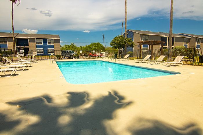 Inviting apartment complex with a sparkling outdoor swimming pool, surrounded by lounge chairs and palm trees, under a clear blue sky.