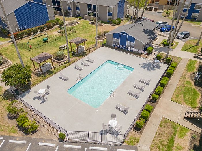 Inviting aerial view of a modern apartment complex, showcasing a pristine swimming pool with surrounding lounge chairs, cozy gazebos, and lush green landscaping for relaxation and leisure.