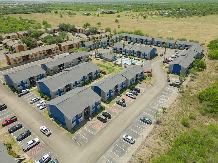 Aerial view of a modern apartment complex surrounded by lush greenery, featuring multiple two-story buildings, ample parking spaces, and a central courtyard with a pool.