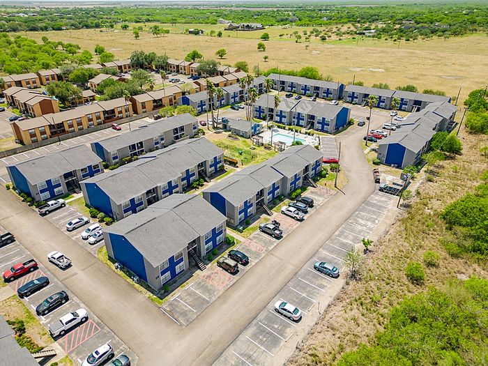 Aerial view of a charming apartment complex surrounded by lush greenery, featuring modern buildings and ample parking space, nestled in a tranquil suburban setting.