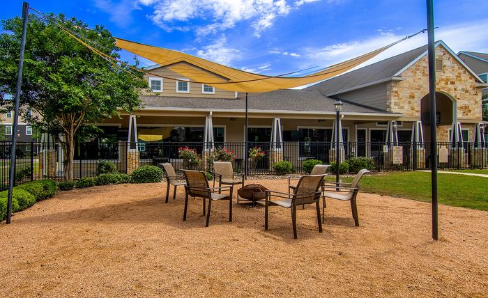 Charming outdoor seating area with modern chairs and a fire pit, framed by lush greenery and a stylish building backdrop, under a vibrant blue sky.