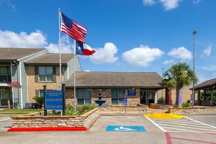 Inviting entrance to Westwood Village Apartments with lush landscaping, vibrant flags, and a welcoming leasing center under a bright blue sky.