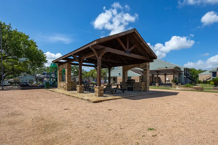 Charming outdoor pavilion with picnic tables and a barbecue grill, set in a vibrant community with a playground and landscaped grounds under a bright blue sky.