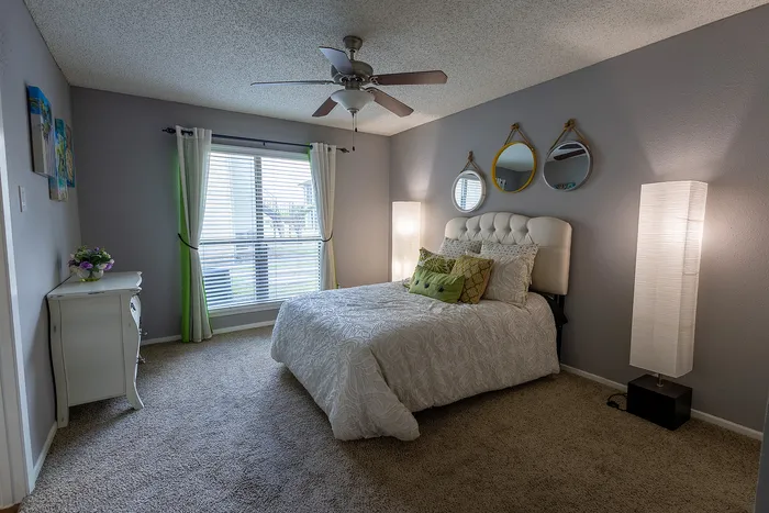 Elegant bedroom with plush carpeting, stylish decor, and abundant natural light from large window. Features a cozy bed adorned with accent pillows and modern lighting.