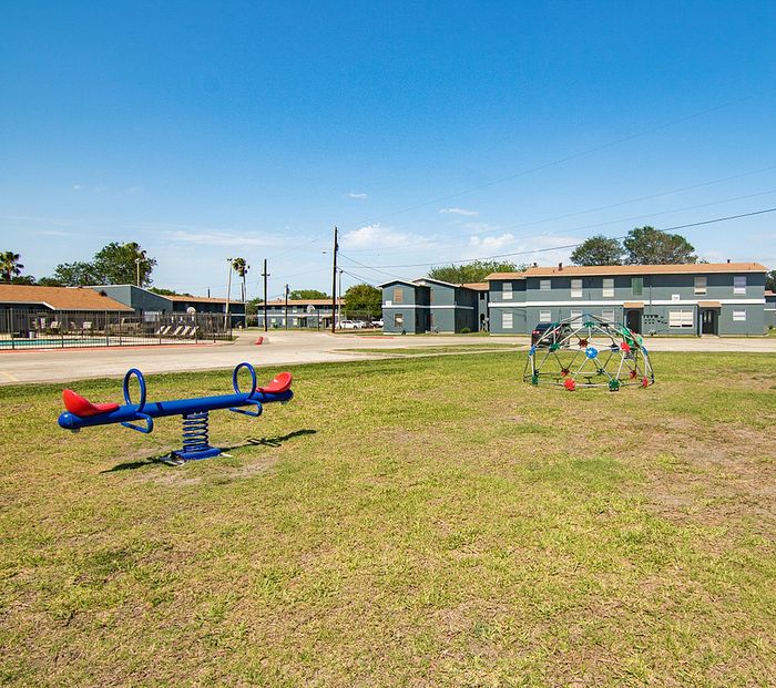 Inviting community playground with see-saw and climbing dome set against modern apartment buildings under a bright blue sky.