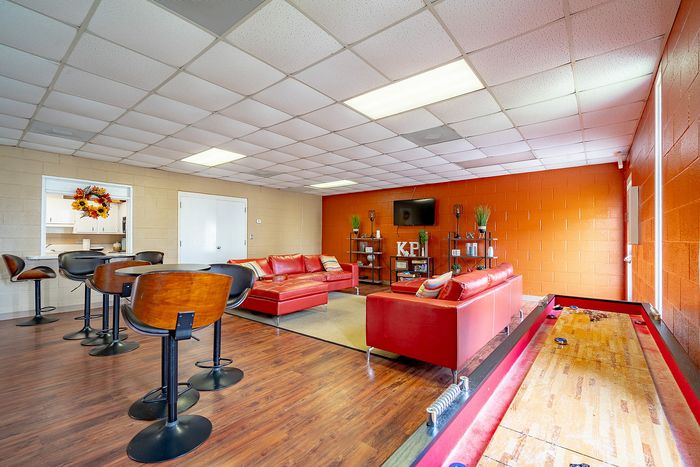 Modern lounge area featuring vibrant red sofas, wooden bar stools, and a shuffleboard table, perfect for relaxation and entertainment.
