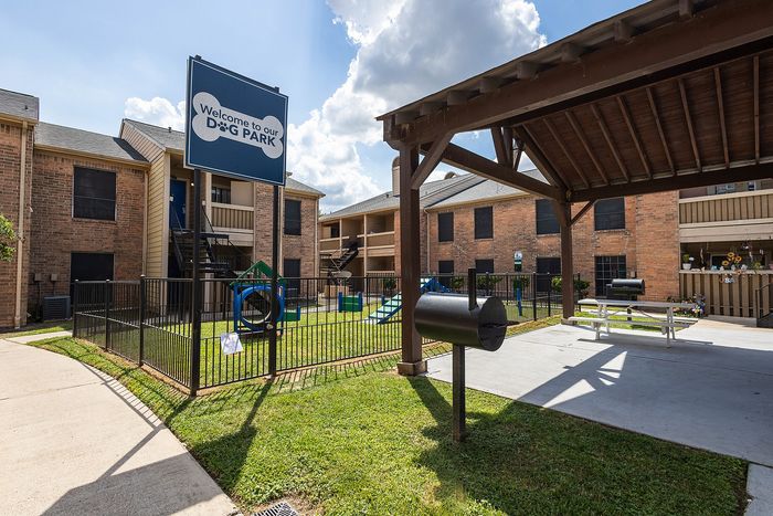 Charming apartment complex featuring a welcoming dog park with playful structures under sunny skies, ideal for pet-friendly living.
