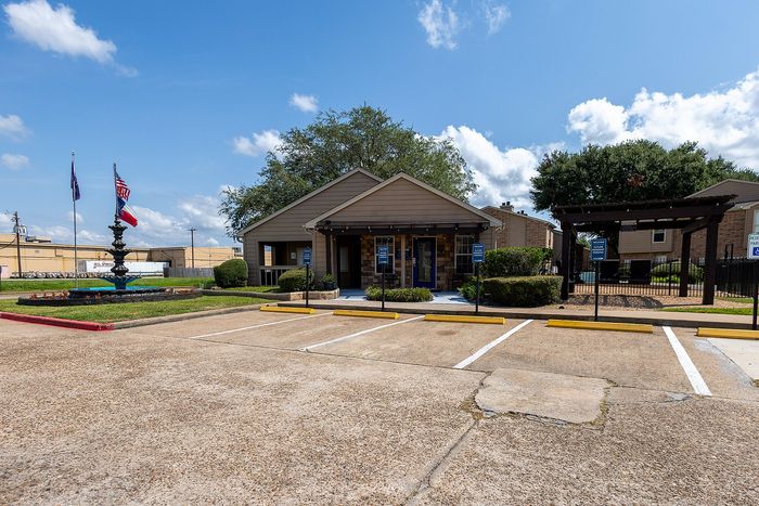 Charming apartment complex entrance with American flag, lush landscaping, and ample parking under a bright blue sky.