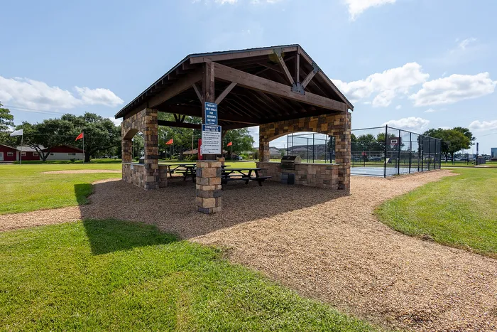 Inviting outdoor pavilion with BBQ grill and picnic tables next to a fenced sports court, surrounded by lush greenery and open skies.