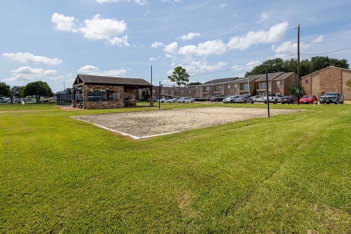 Inviting outdoor area featuring a sand volleyball court, vibrant green lawn, and charming pavilion shelter with modern apartment buildings in the background.