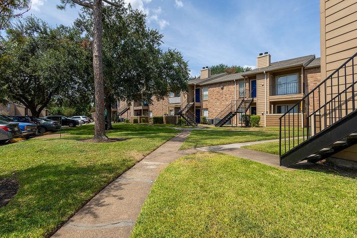 Charming apartment complex with lush green lawns, mature trees, and inviting brick facades, ideal for serene living.