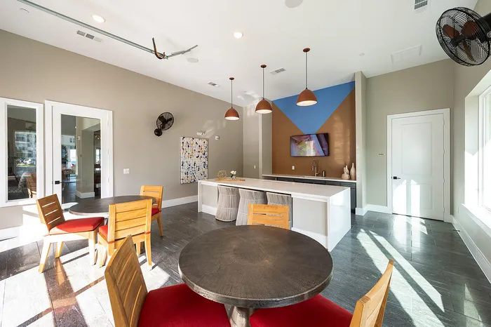 Contemporary communal area with chic round tables, vibrant red chairs, and a sleek kitchen space with stylish pendant lighting, perfect for social gatherings.