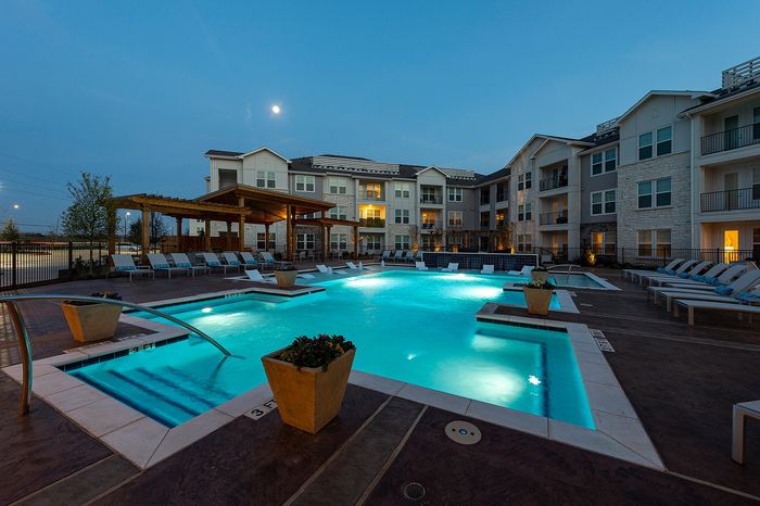 Luxurious apartment complex with a beautifully illuminated swimming pool and ample lounge chairs at dusk.