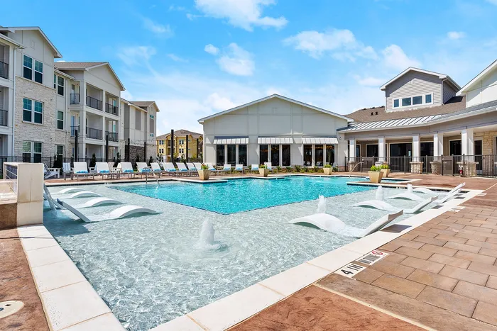 Luxury apartment complex featuring a stunning outdoor pool, modern lounge chairs, and beautifully landscaped surroundings under a bright blue sky.