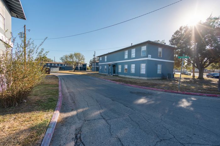 Charming residential neighborhood with modern two-story blue apartment buildings, surrounded by lush greenery and bathed in warm sunshine.
