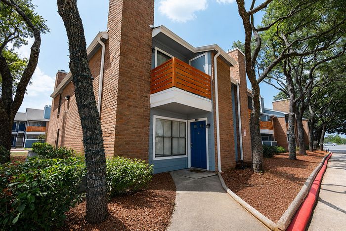 Exterior of Deerbrook Gardens apartments in Humble, TX featuring brick buildings, mature trees and updated wood balcony enclosures.