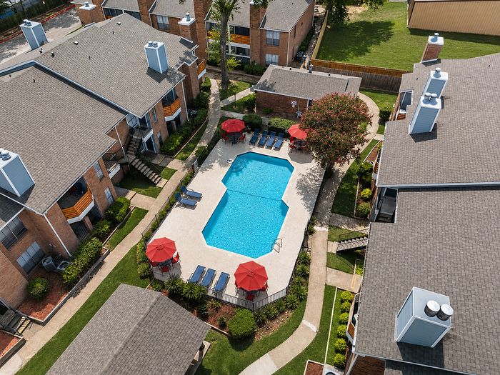 Swimming pool at Deerbrook Gardens in Humble, TX with red lounge seating, umbrellas and surrounding apartment buildings.
