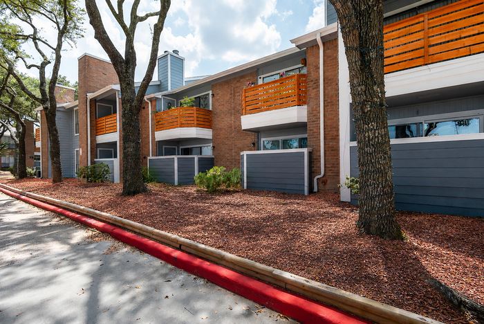 Tree-lined apartment exterior at Deerbrook Gardens in Humble, TX with landscaped beds, brick architecture and private balconies.
