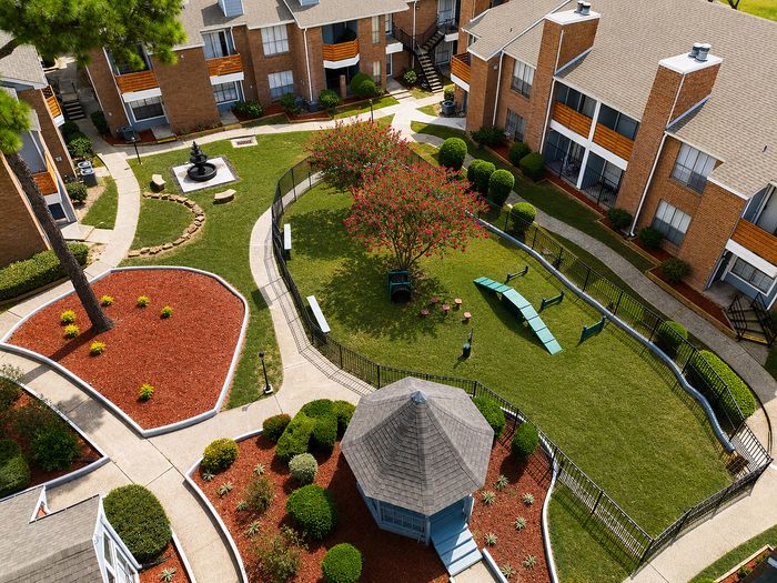 Aerial view of the Deerbrook Gardens dog park in Humble, TX featuring agility equipment, a flowering shade tree and surrounding courtyard.