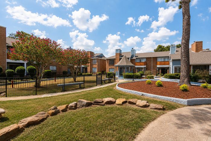 Fenced dog park at Deerbrook Gardens in Humble, TX with agility equipment, shade trees, benches and landscaped surroundings.