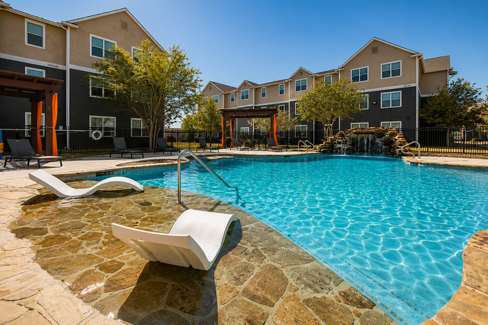 Luxurious apartment complex featuring a pristine swimming pool with a waterfall, lounge chairs, and landscaped surroundings.