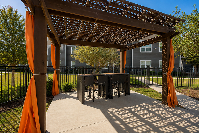 Charming outdoor patio with elegant wooden pergola, vibrant orange curtains, and stylish bar seating, nestled in a serene, landscaped garden setting.
