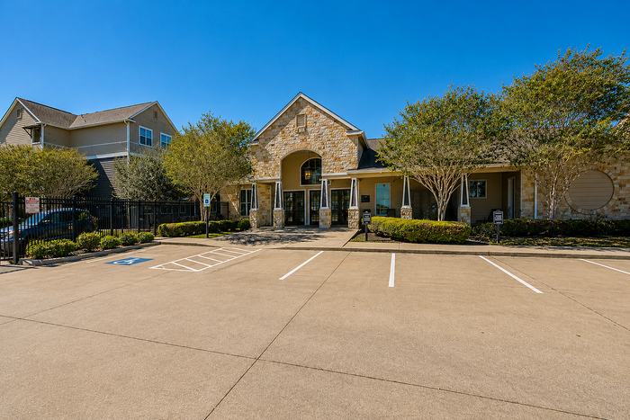 Charming apartment complex entrance with stone accents, landscaped greenery, and ample parking under a clear blue sky.