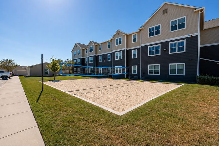 Inviting apartment complex with a well-maintained sand volleyball court, surrounded by lush greenery and clear blue skies, offering an active lifestyle.