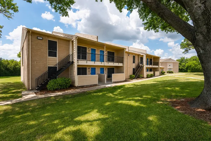Exterior of The Reserve on Garth apartments in Baytown, TX featuring beige brick buildings, blue doors, mature trees and landscaped lawns.
