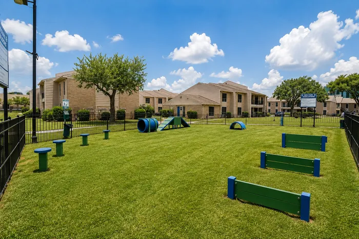 Fenced dog park with green and blue agility equipment and open lawn at The Reserve on Garth apartments in Baytown, TX.