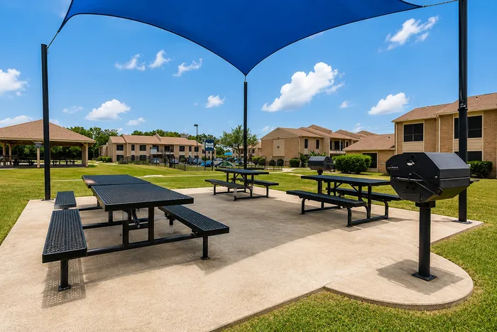 Outdoor grilling and picnic area beneath a blue shade canopy with the community pool visible in the background at The Reserve on Garth.