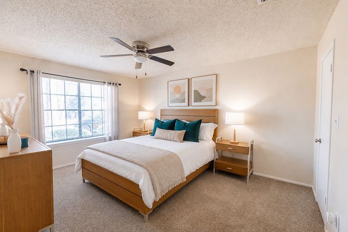 Furnished bedroom at Apple Creek apartments with a large window, bedside tables, ceiling fan and neutral finishes.