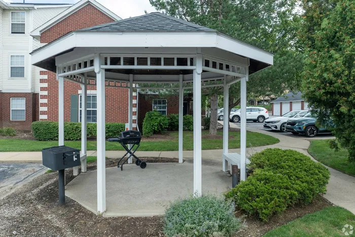 Charming outdoor gazebo with a convenient barbecue grill, set amidst lush greenery and elegant brick architecture.